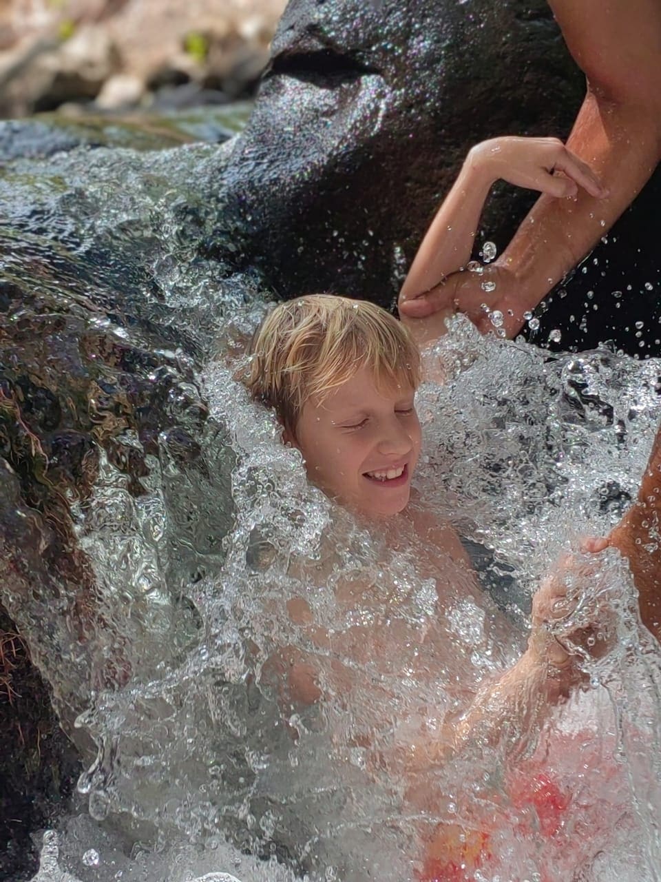 Enfant sourire, joue dans l'eau de cascade, soutenu par un adulte.