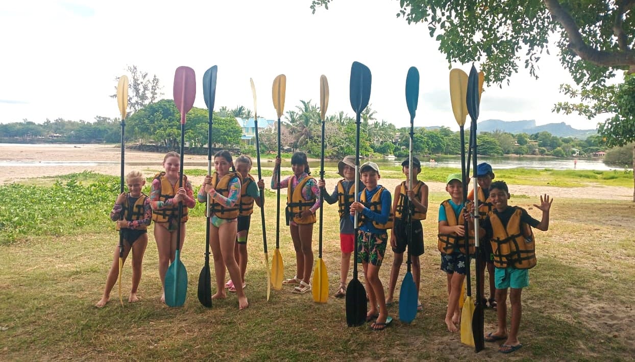 Enfants en gilets de sauvetage avec pagaies colorées sur pelouse près d'un plan d'eau.