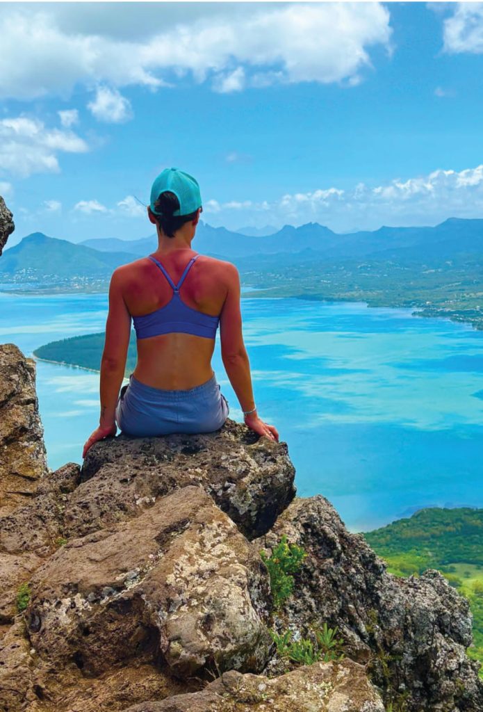 Personne assise dos à la caméra sur rochers face à un lac turquoise et montagnes sous ciel nuageux.